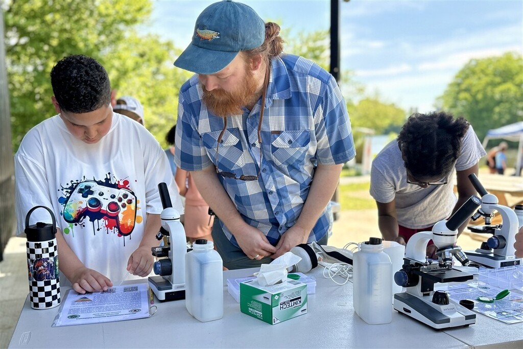 A group of people, including children and an adult, gather around a table with microscopes and scientific equipment outdoors.