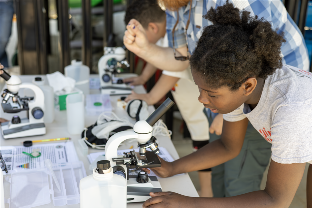 A young person intently examines a specimen under a microscope at an outdoor science station.