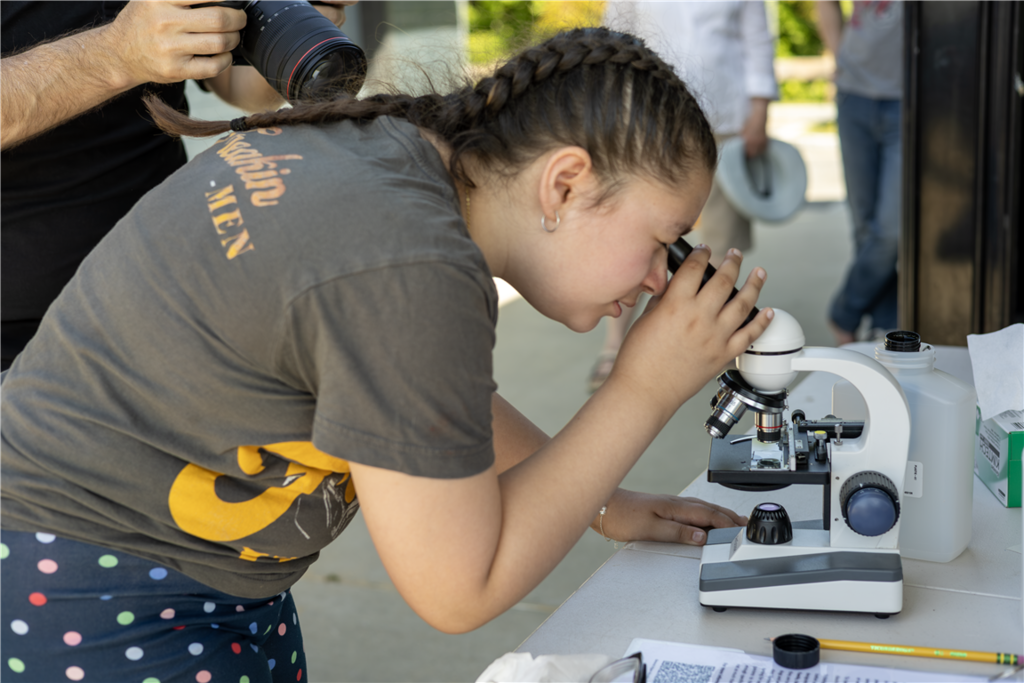 A young person with braided hair peers through a microscope on a table.