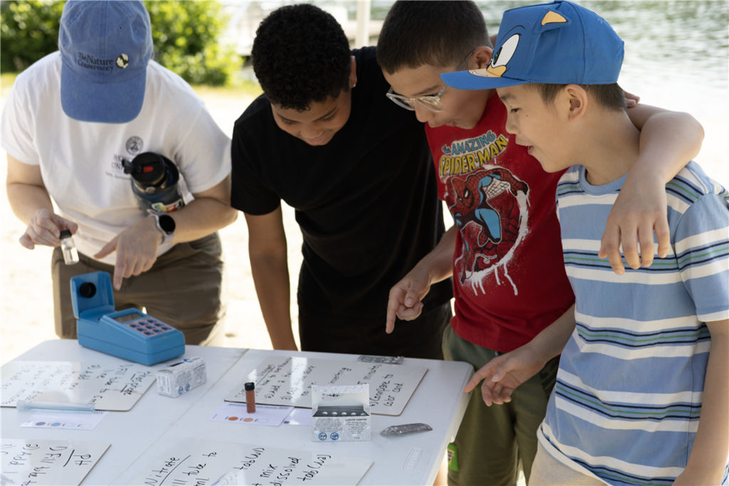 A group of children and an adult examine a water testing kit on a table outdoors.