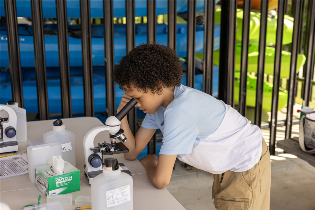 A young boy with curly hair peers intently into a microscope on a table.