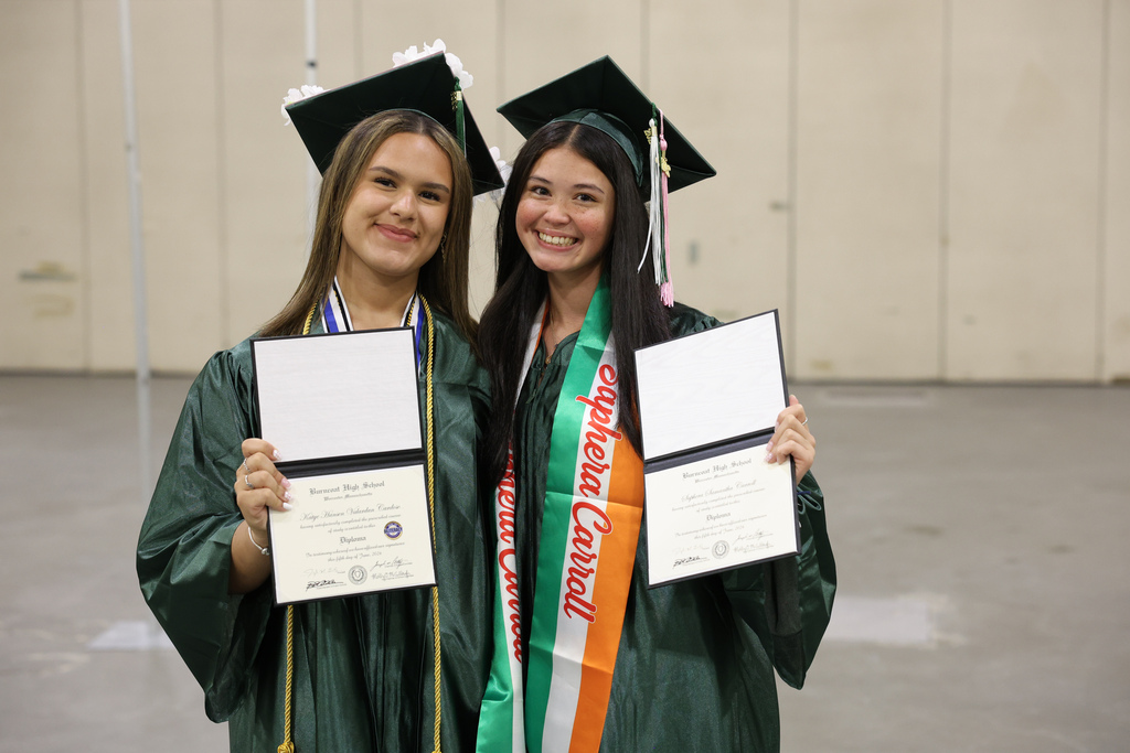 Two students hold up their high school diplomas while wearing caps and gowns.