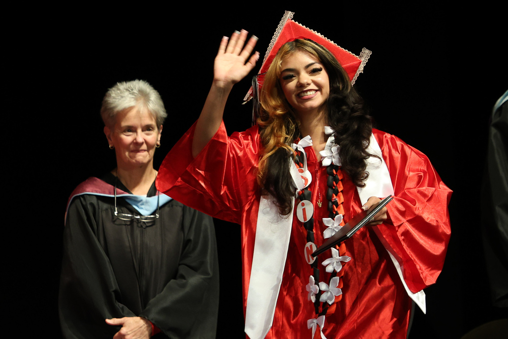 A student smiles and waves as they walk across stage at graduation.