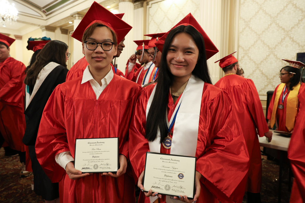 Two high school students in caps and gowns hold their high school diplomas.