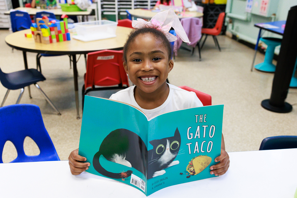 A student smiles wide while holding up their book called "The Gato Taco".