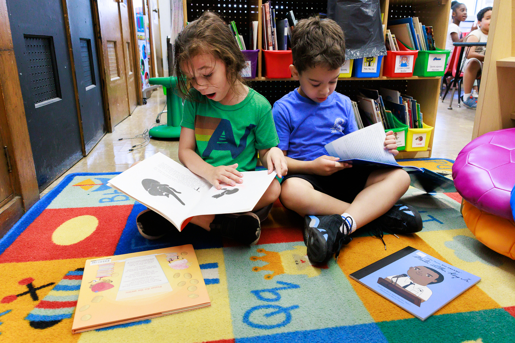 Two students sit on a carpet and flip through books.
