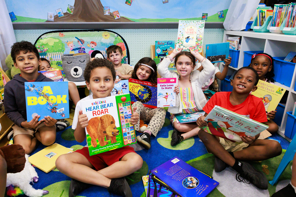Students sit on the class rug and hold up their books.