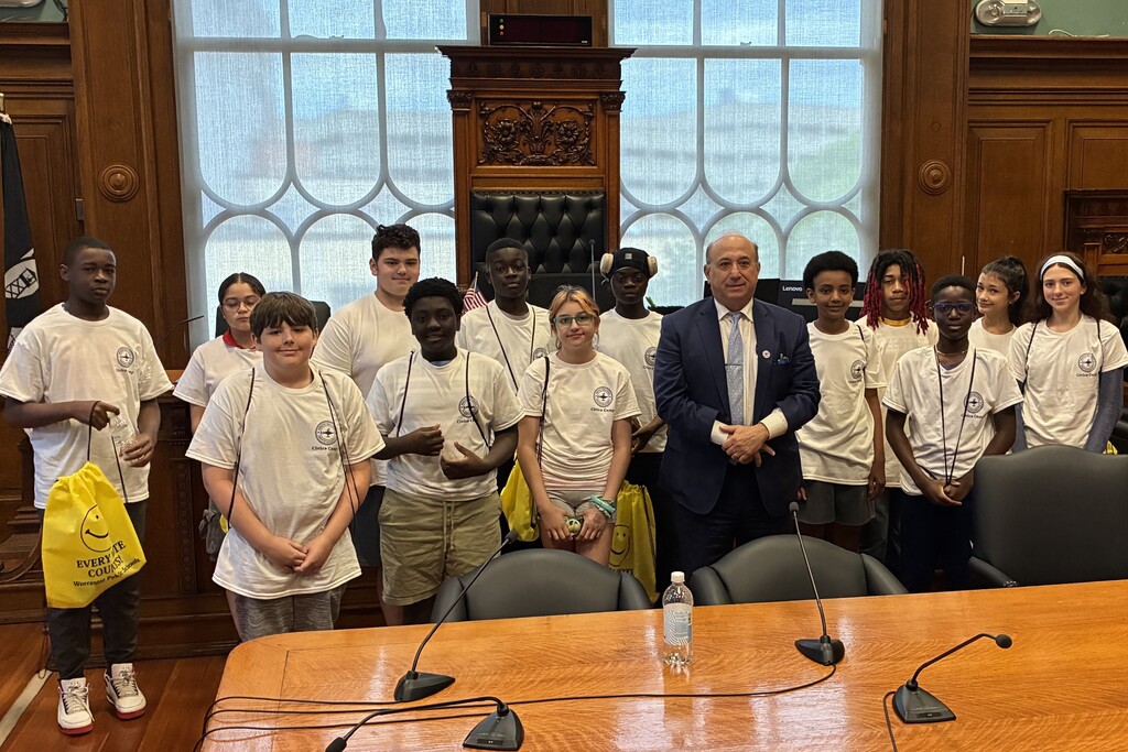 A group of young people in matching t-shirts stand with a man in a suit in a room with ornate wooden paneling and large windows.