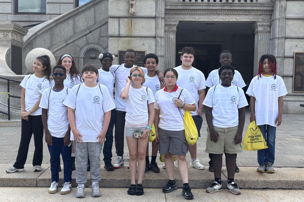 A diverse group of young people in matching white t-shirts stand on stone steps in front of a building.