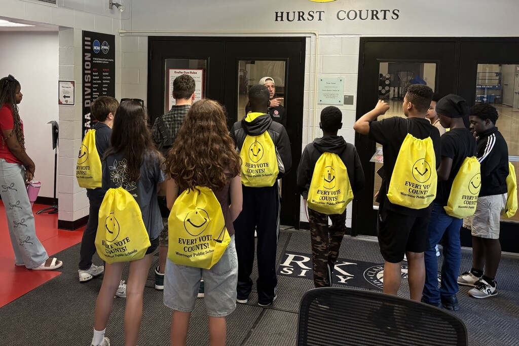 A group of students wearing yellow backpacks with the slogan 'EVERY VOTE COUNTS!' stand near a sign for 'HURST COURTS'.