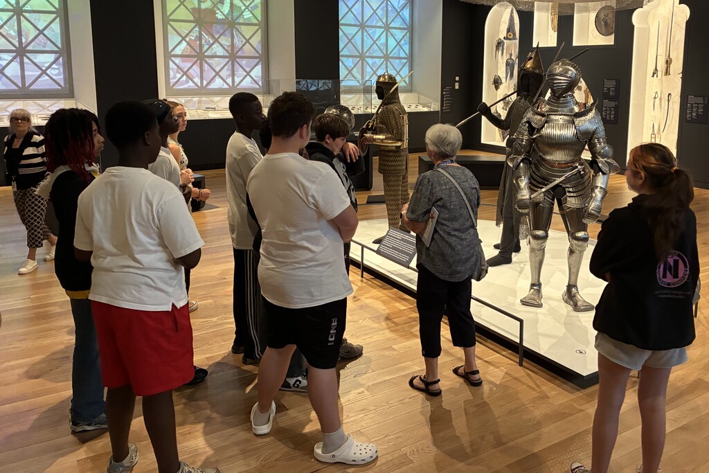 A group of young people stand in a museum exhibit featuring suits of armor and historical weapons.
