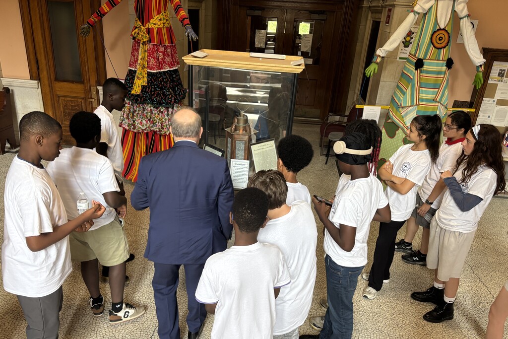 A group of students in white t-shirts listen to a man in a suit in front of a display case and large colorful figures.