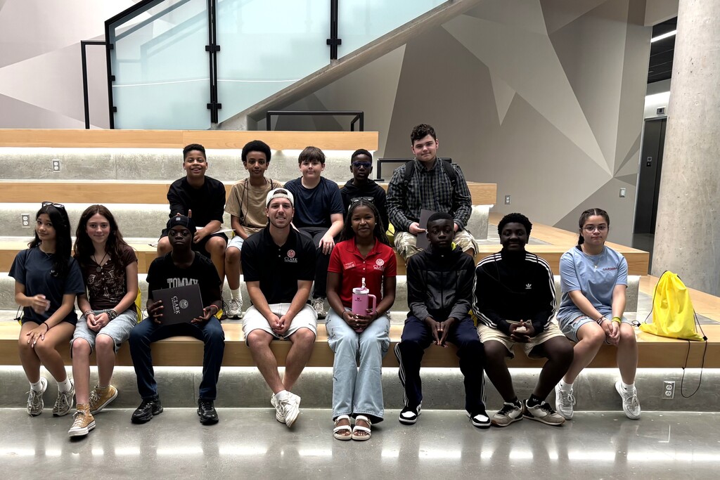 A diverse group of young people and an adult sit on tiered wooden benches in a modern building.
