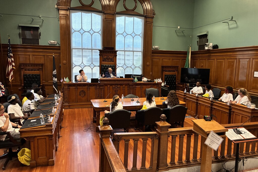 A group of young people sit at desks in a courtroom, facing a judge and another individual at the front.