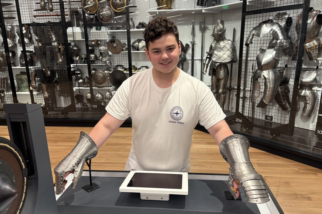 A young person wearing a white t-shirt with a 'Civics Camp' logo smiles while trying on ornate metal gauntlets in a museum exhibit.