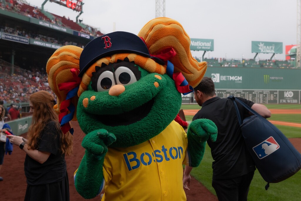 A green mascot with a red 'B' hat and a yellow 'Boston' jersey stands on a baseball field.