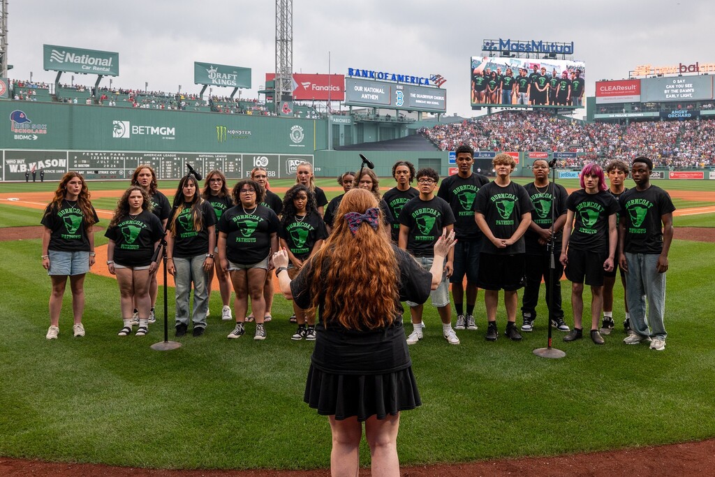 A choir of students in matching 'Burncoat Patriots' t-shirts stands on a baseball field, facing a crowd.