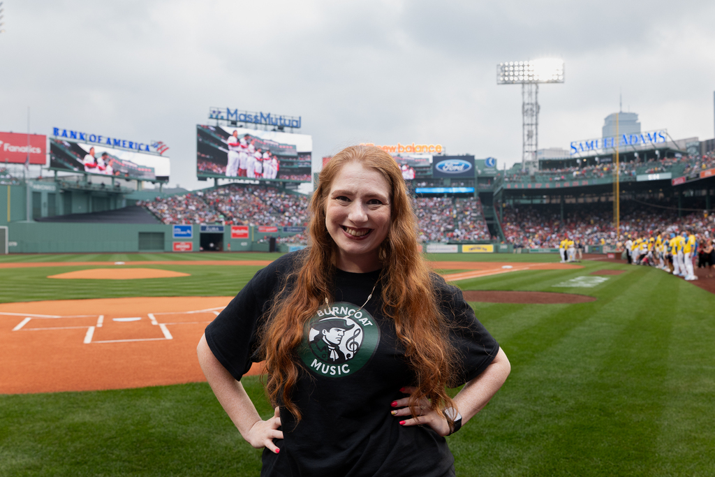 A woman with long red hair smiles at the camera on a baseball field, wearing a black t-shirt with a circular logo.