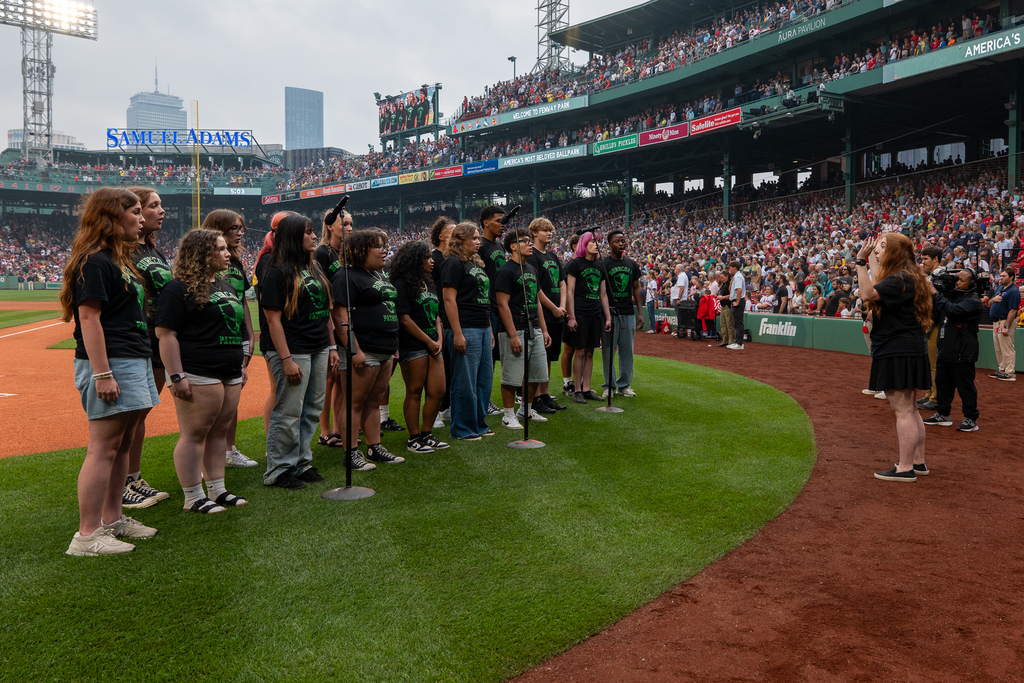 A group of young people in matching t-shirts stand on a baseball field, facing a crowd in the stands.