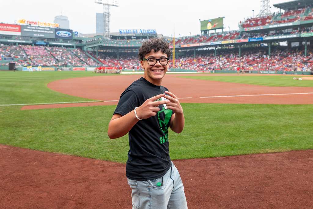 A smiling young person stands on a baseball field, with stadium seating and a crowd in the background.