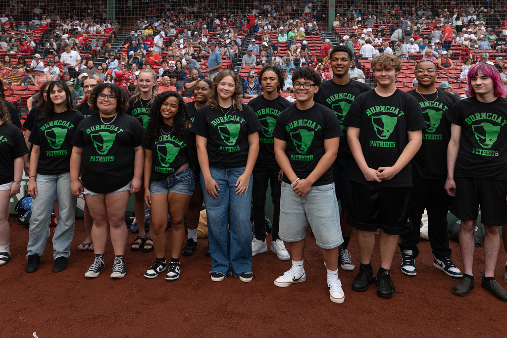 A diverse group of young people wearing matching black "Burncoat Patriots" t-shirts stand on a baseball field