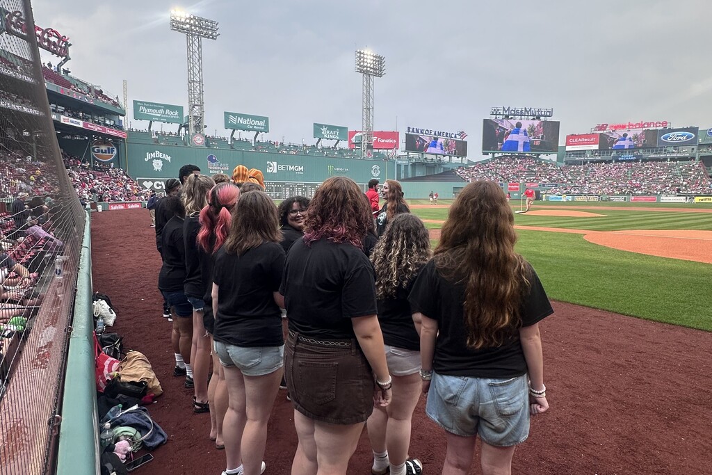 A group of people stand on a baseball field, facing away from the camera, with stadium seating and advertisements visible in the background.