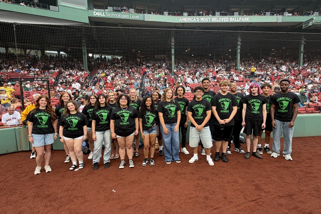 A group of young people wearing matching black t-shirts with a green logo stand on a baseball field.