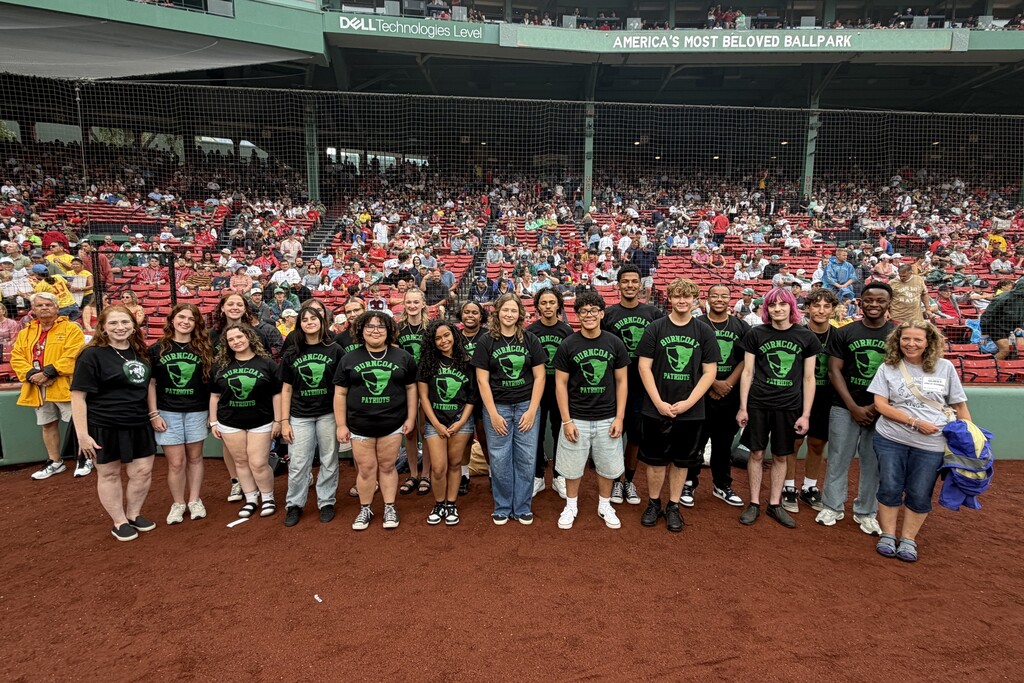 A group of young people wearing matching black t-shirts with a green logo stand on a baseball field.