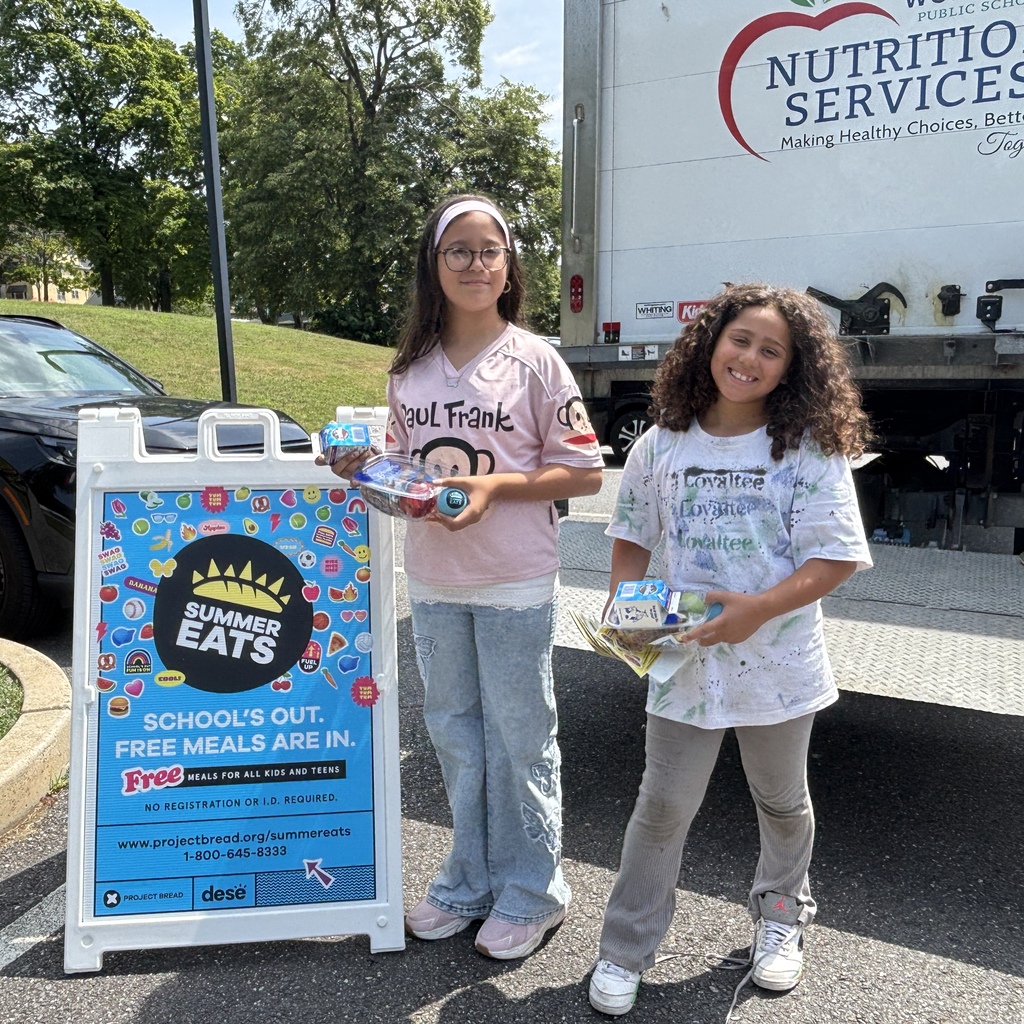 Two young girls stand next to a sign advertising "Summer Eats" free meals, holding food packages.