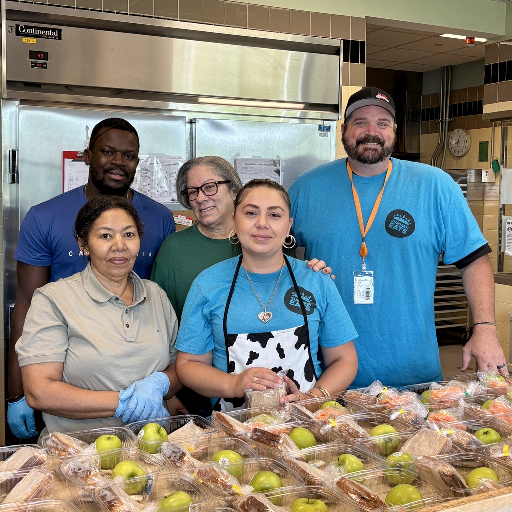 Five smiling individuals stand behind a table laden with pre-packaged meals, including sandwiches and apples.