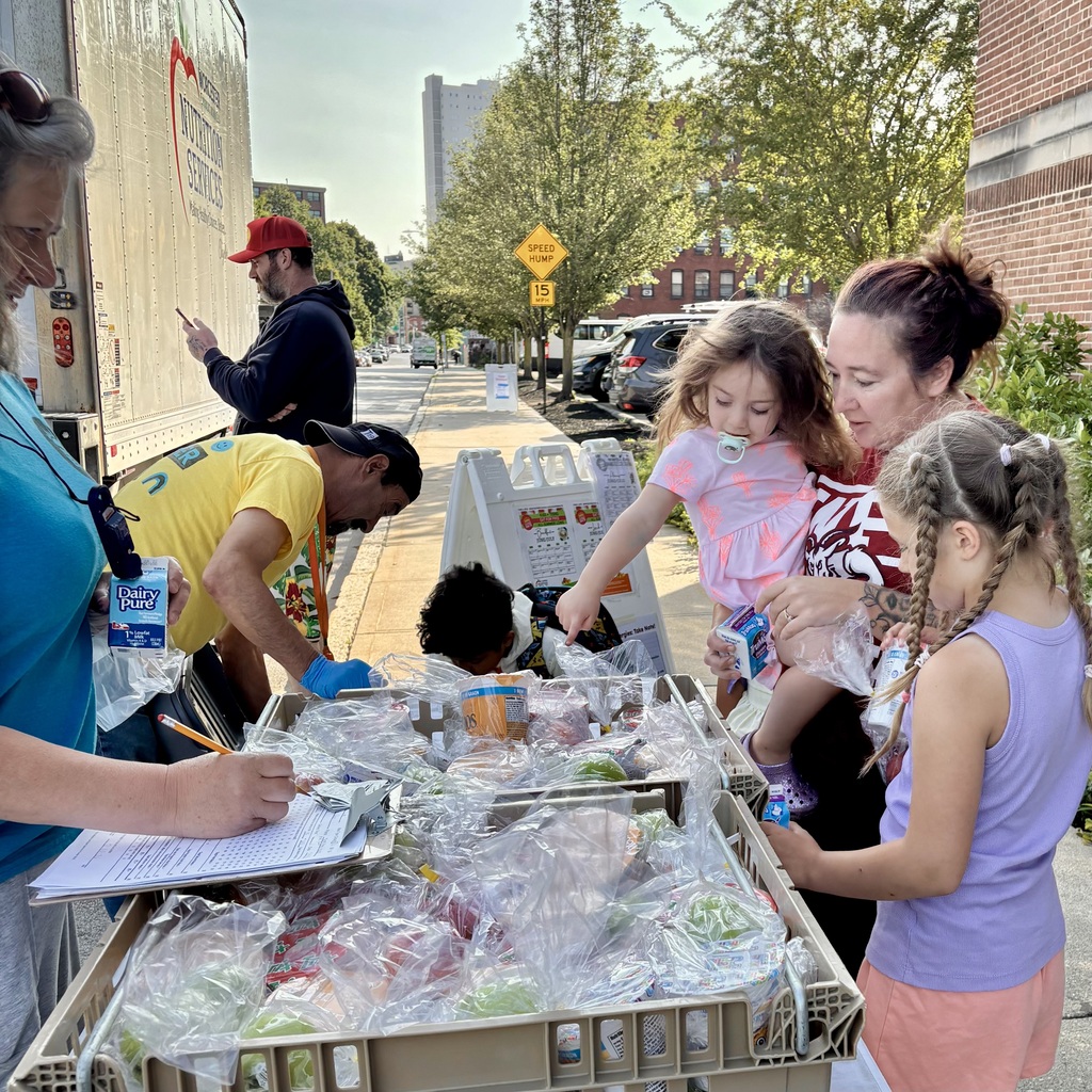 Nutrition Services staff distribute pre-packaged meals from a truck labeled “Worcester Public Schools Nutrition Services” to a family on a sidewalk.