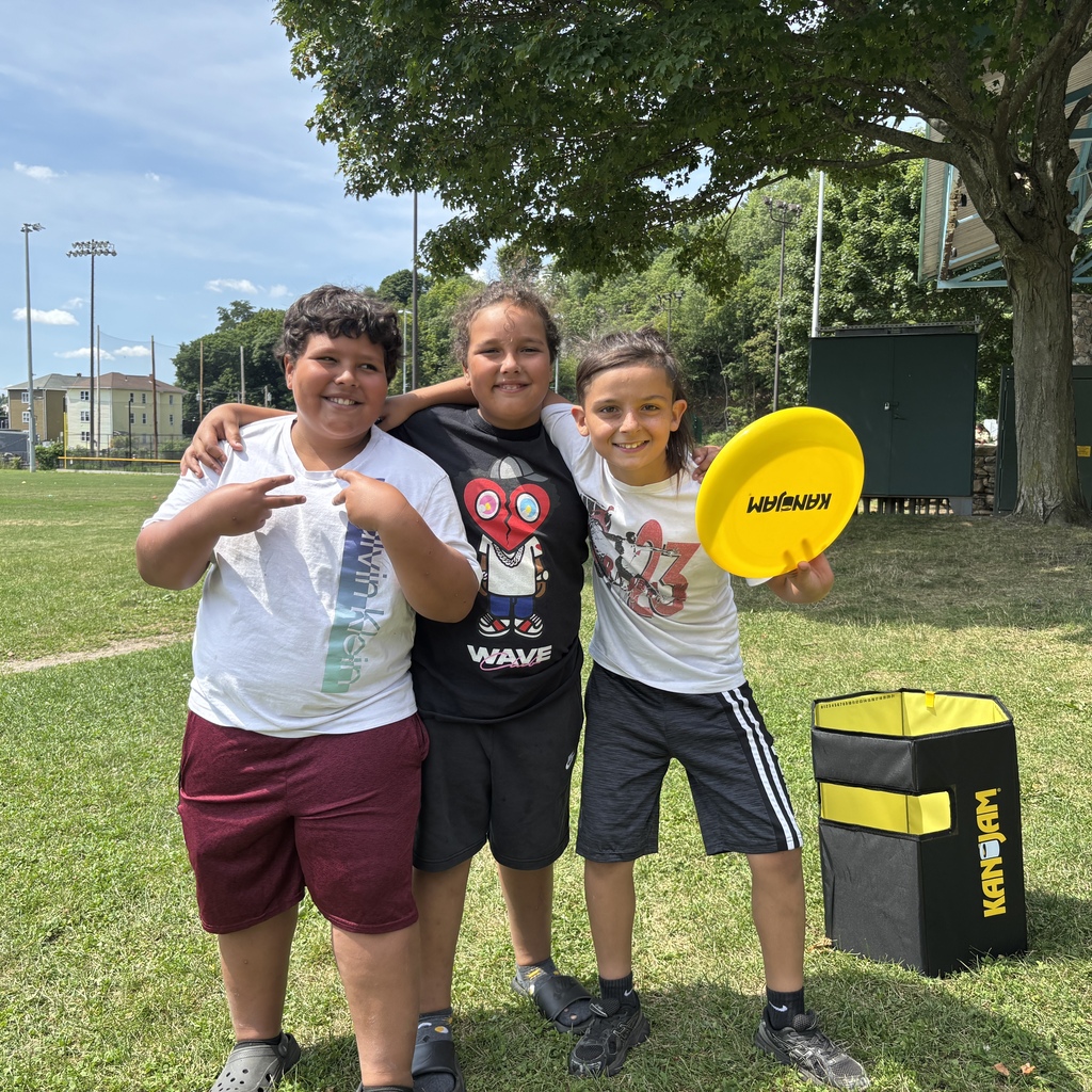 Three smiling children pose outdoors with a yellow KanJam disc and game set.