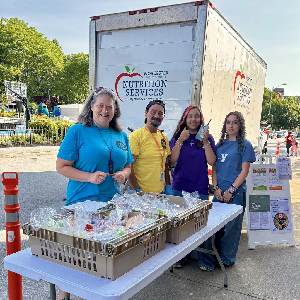 Four people stand behind a table laden with bagged food items, with a large truck labeled 'Worcester Public Schools Nutrition Services' in the background.