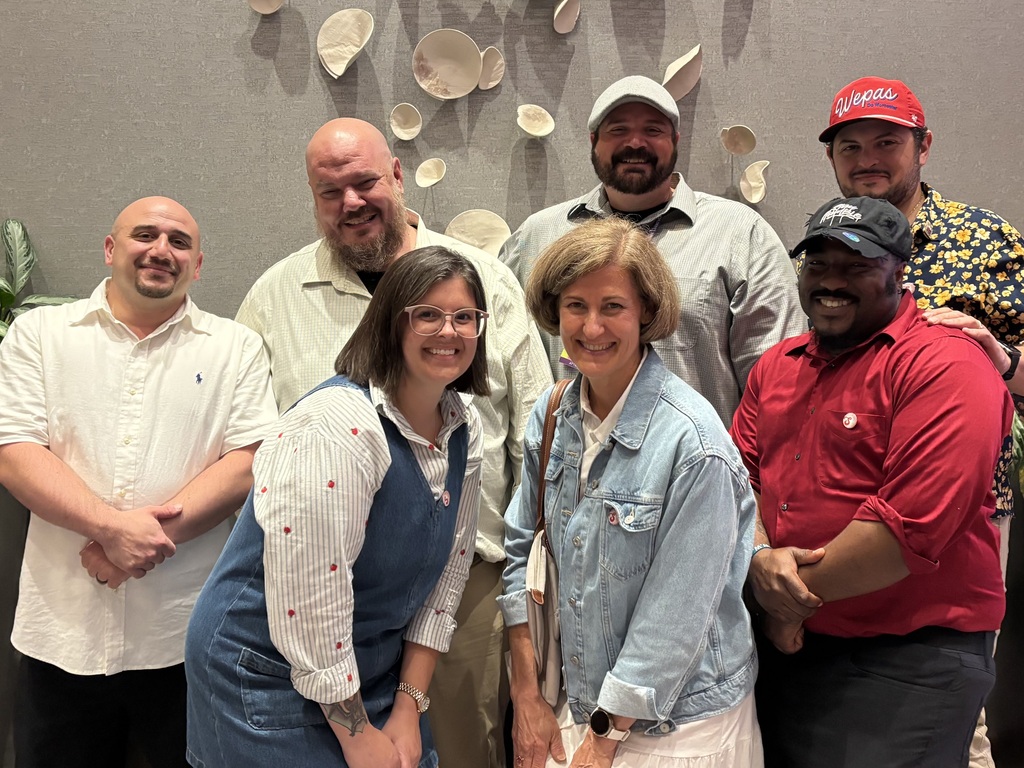 A diverse group of seven people smile for a photo in front of a textured wall with decorative ceramic pieces.