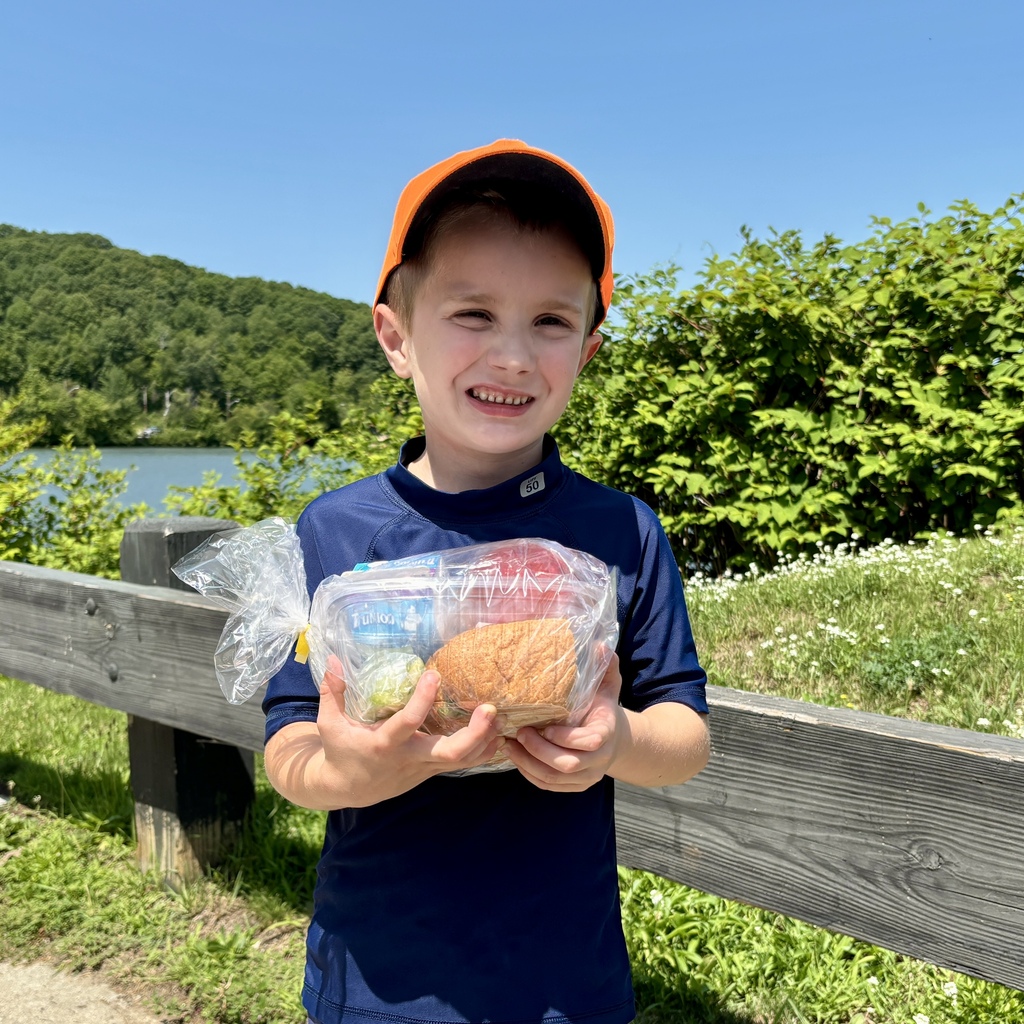 A smiling boy in an orange hat holds a clear plastic bag containing a sandwich and drink.