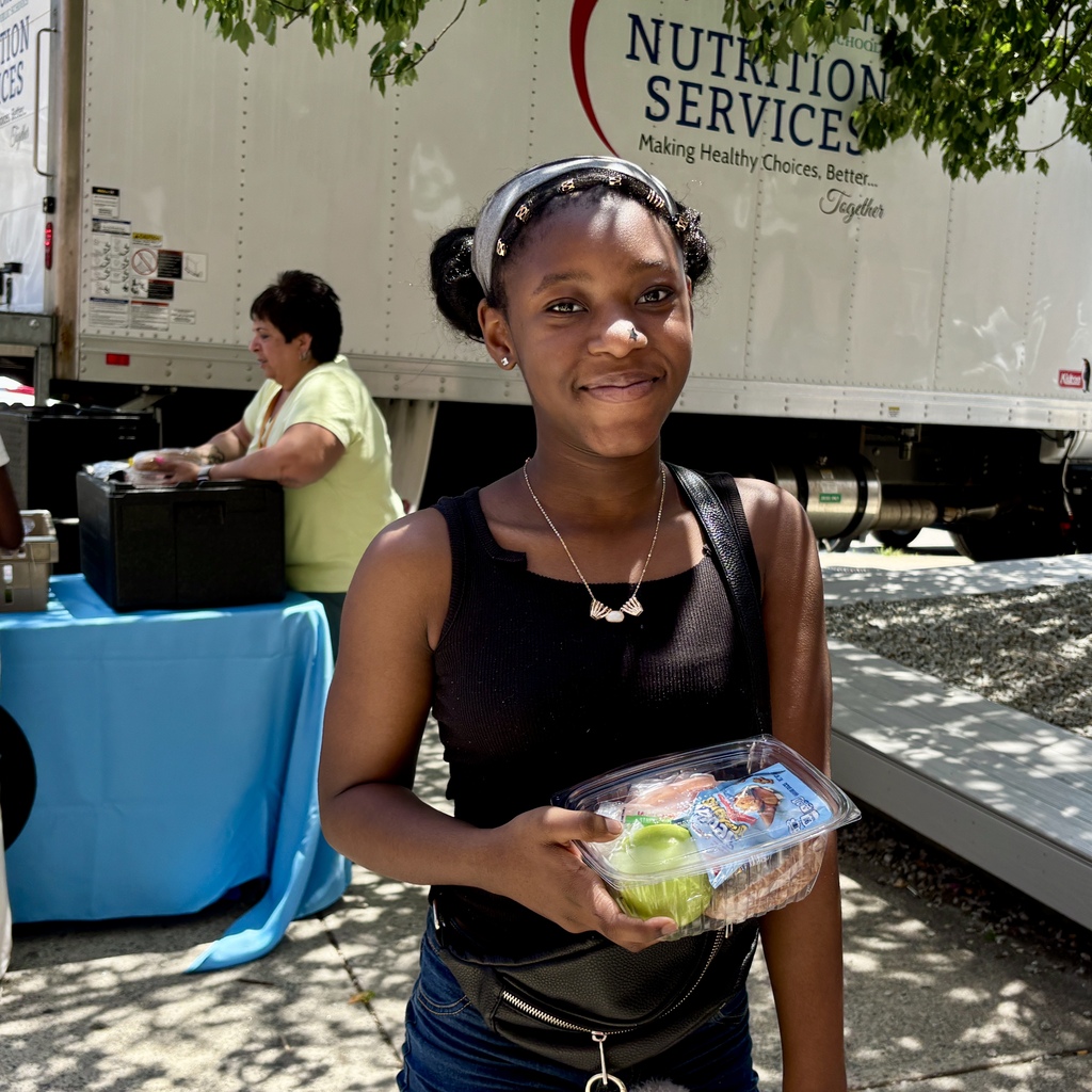 A young woman smiles while holding a container with a green apple and a sandwich.