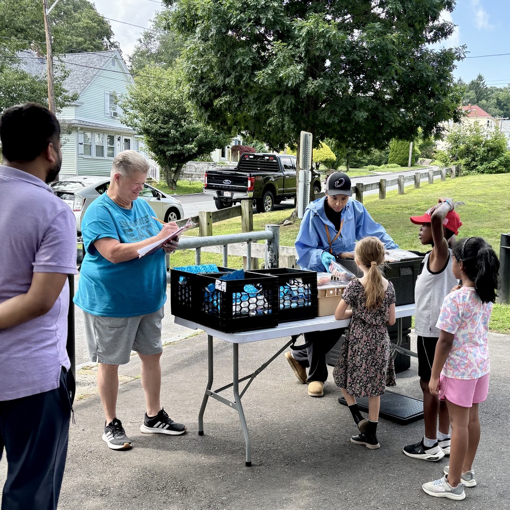Volunteers distribute food items from crates on a table to children and adults outdoors.