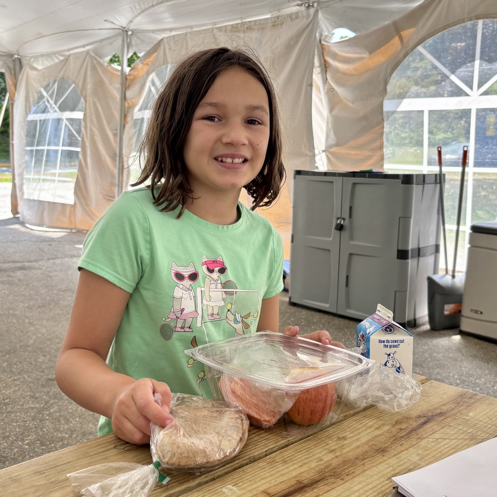 A young girl in a green t-shirt smiles while sitting at a wooden table with a meal.
