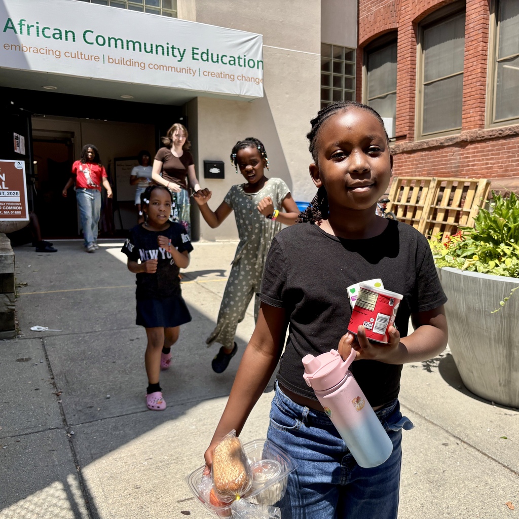 A young girl holds a container of food and a water bottle outside a building with an 'African Community Education' banner.