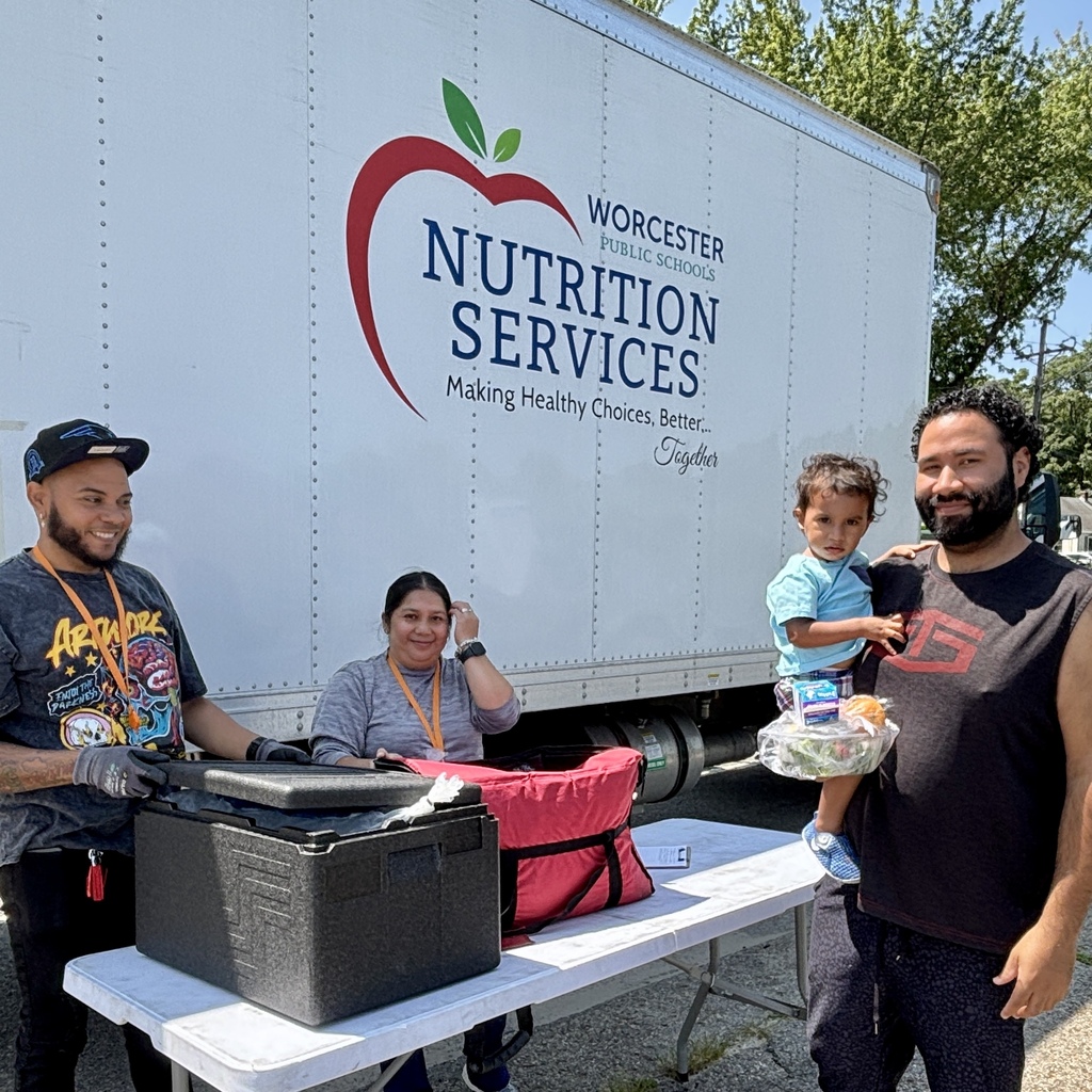 A man holds a child while standing near a table with food distribution supplies, with a truck labeled 'Worcester Public Schools Nutrition Services' in the background.
