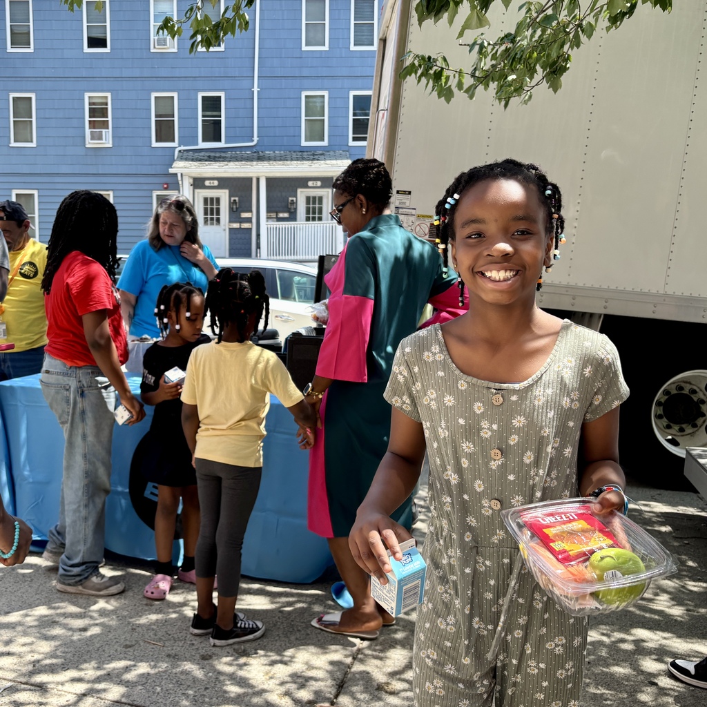 A smiling young girl holds a container of snacks and a carton of milk, with a food distribution event in the background.