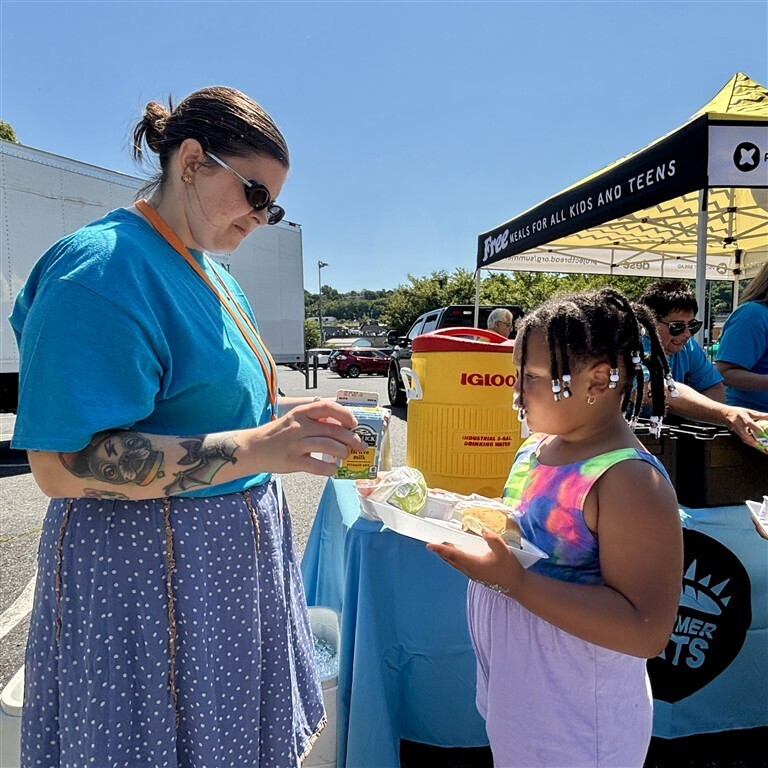 A volunteer hands a child a meal tray at an outdoor event with a tent that reads 'FREE MEALS FOR ALL KIDS AND TEENS'.