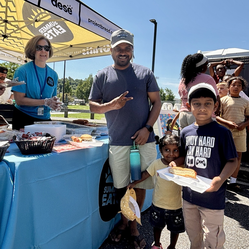 A volunteer hands a child a meal tray at an outdoor event with a tent that reads 'FREE MEALS FOR ALL KIDS AND TEENS'.