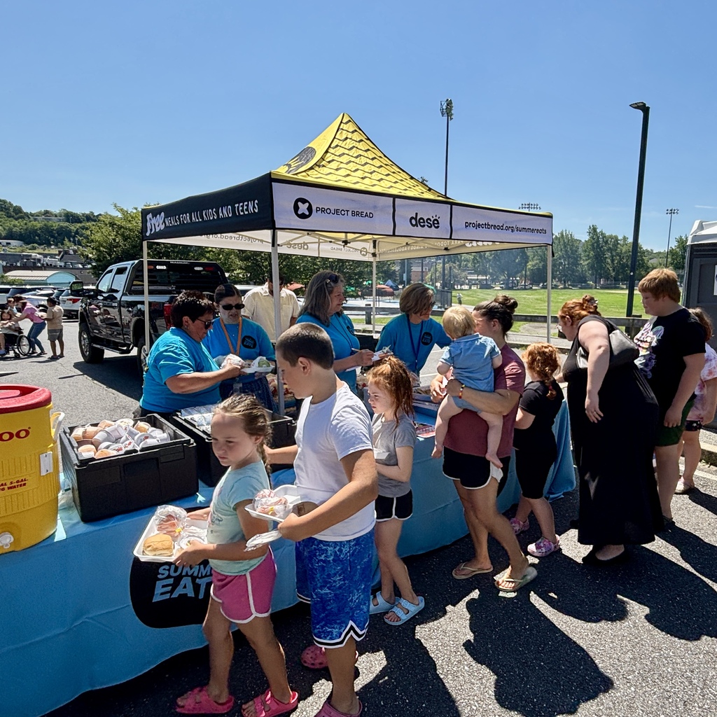 Volunteers distribute free meals for kids and teens under a yellow and black tent at an outdoor event.