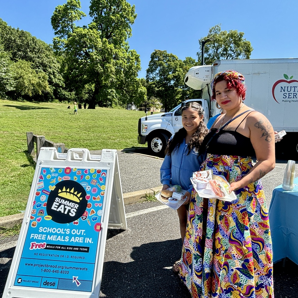 A woman and a girl stand near a sign advertising "Summer Eats" free meals for kids and teens.