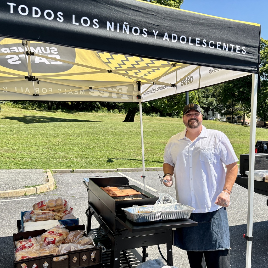 A man in a white shirt and denim apron grills hot dogs under a tent with Spanish text "TODOS LOS NIÑOS Y ADOLESCENTES".