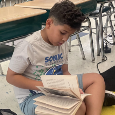 A young boy sits on the floor in a classroom, engrossed in reading a book.