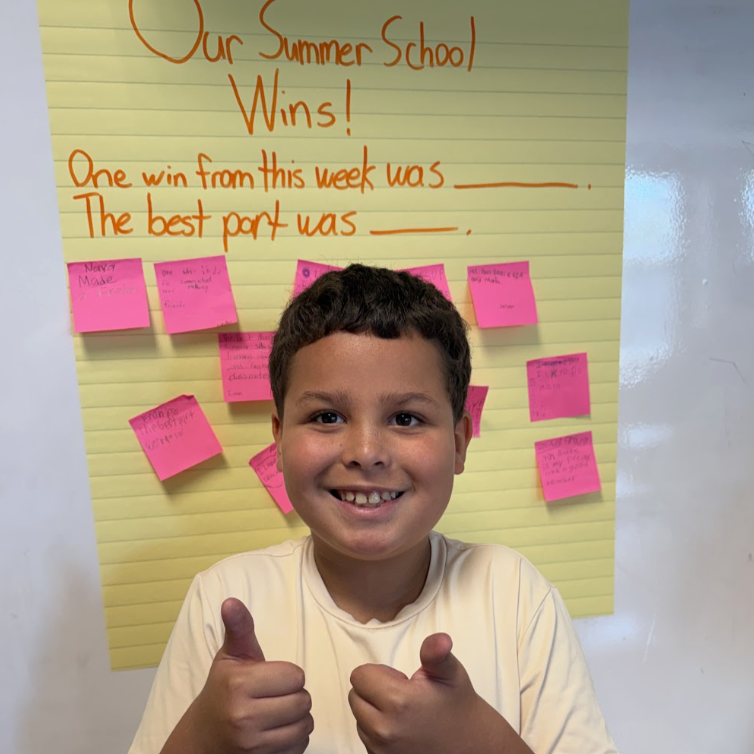 A smiling boy gives two thumbs up in front of a yellow poster board titled 'Our Summer School Wins!'