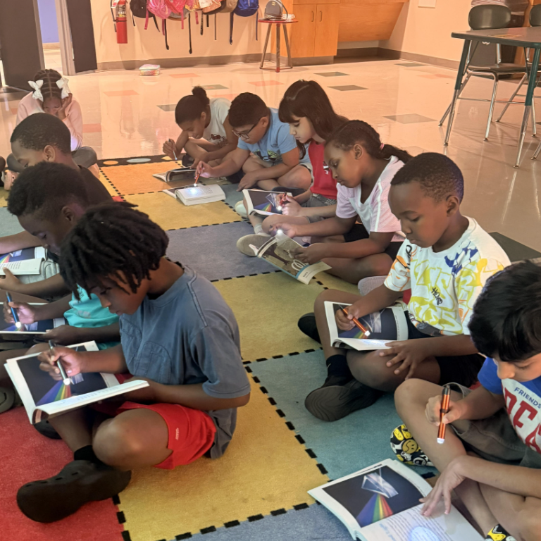 Children sit on a colorful mat, using small flashlights to illuminate their books.