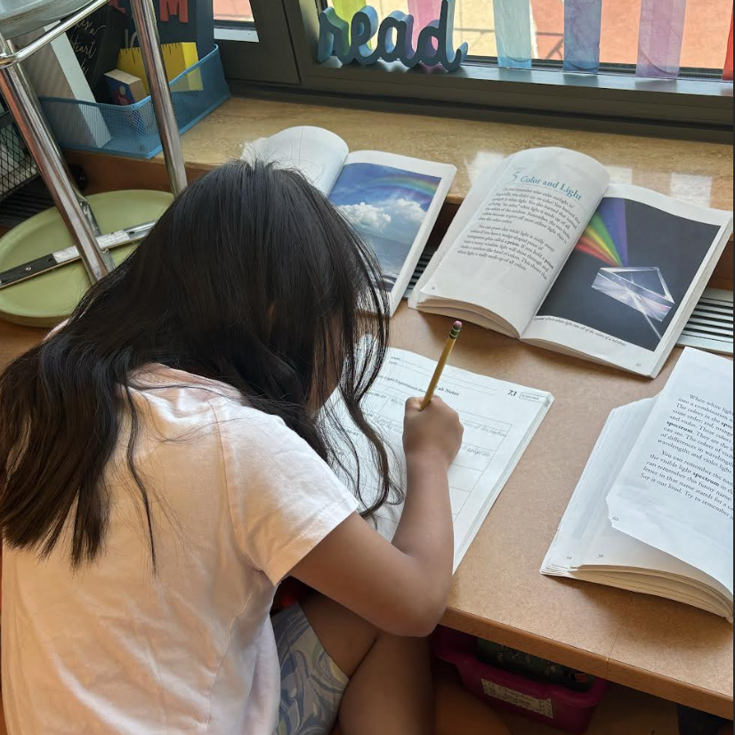 A young student writes in a notebook at a desk with open textbooks about color and light.